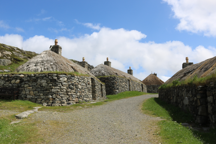 Stone houses with thatched roofs along a gravel path under a blue sky.