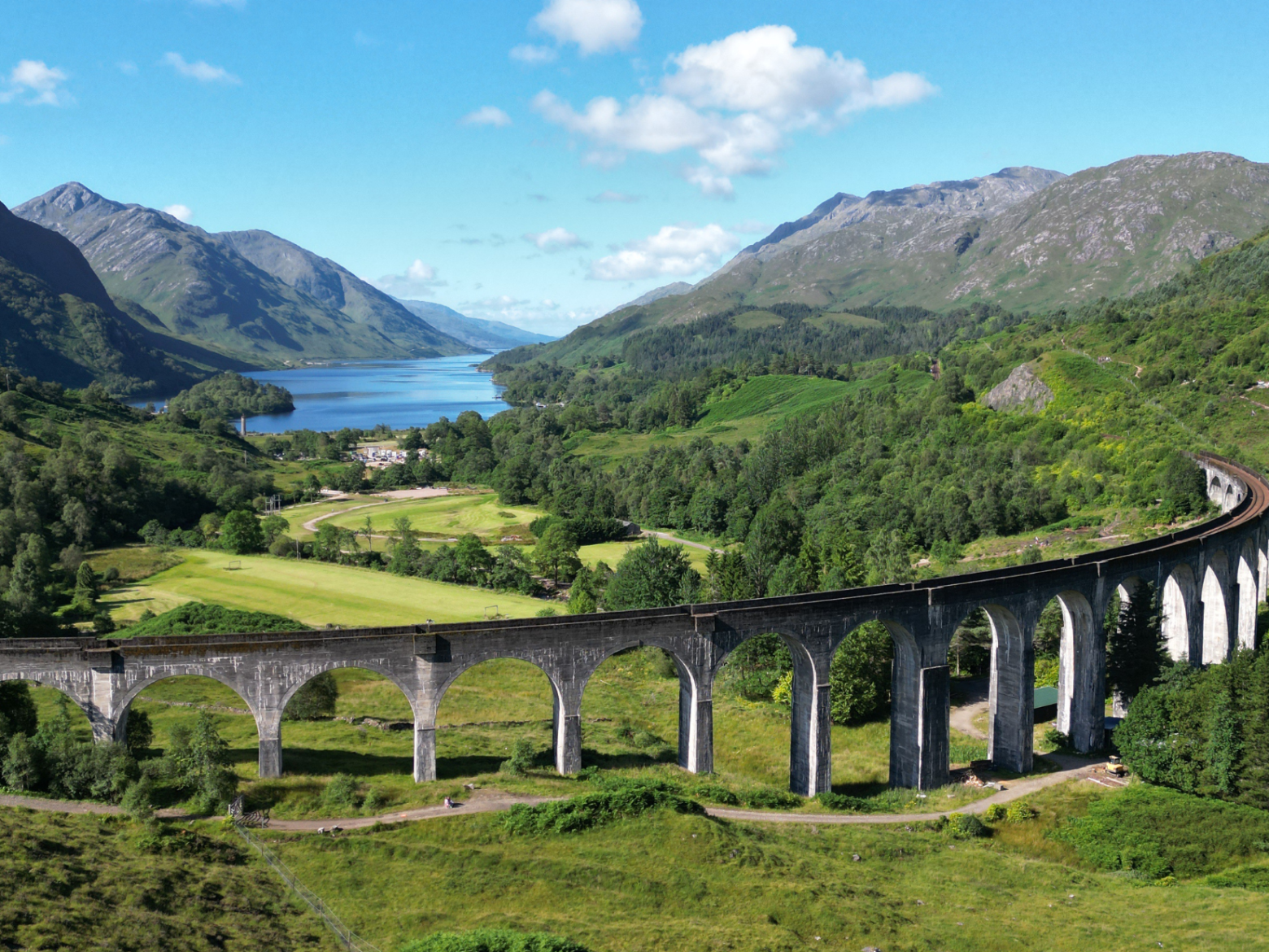 Scenic railway viaduct in lush green valley with mountains and lake in background.