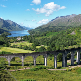 Scenic railway viaduct in lush green valley with mountains and lake in background.