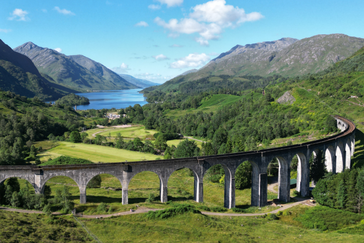 Scenic railway viaduct in lush green valley with mountains and lake in background.