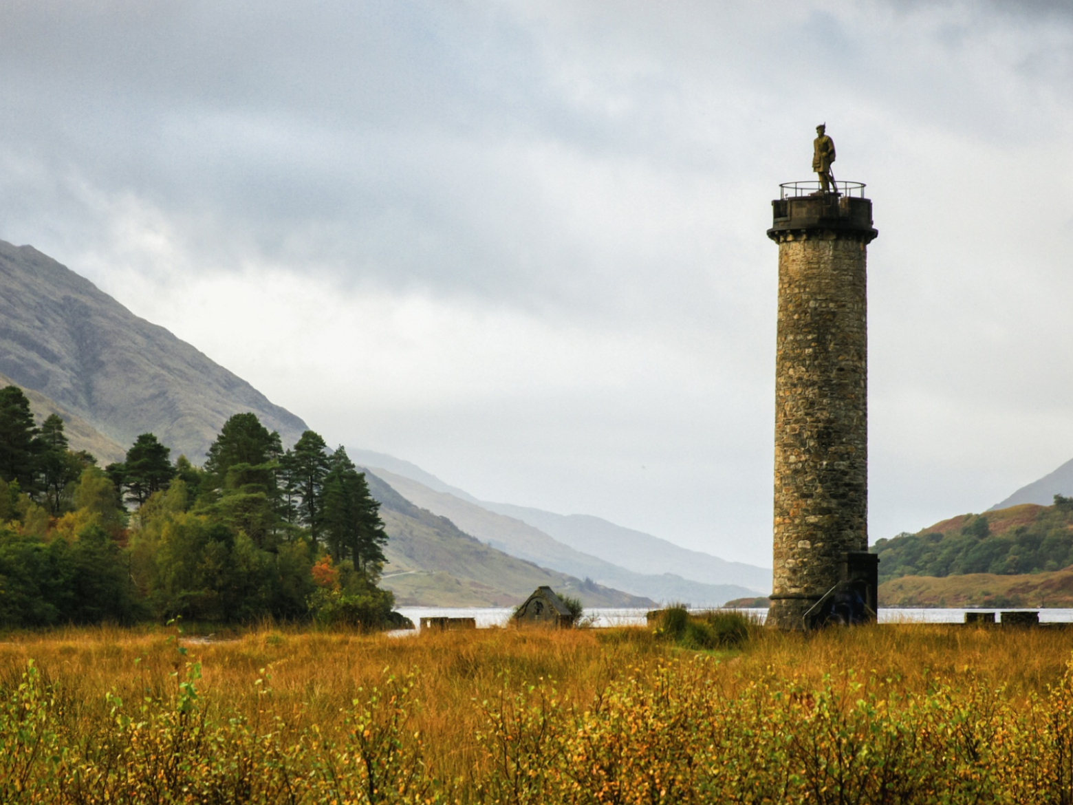 Tall stone tower in a grassy landscape with mountains and a lake.
