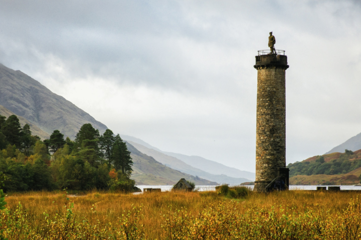 Tall stone tower in a grassy landscape with mountains and a lake.