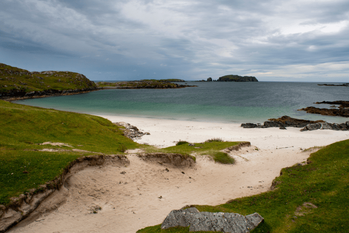 Quiet beach with white sand and green hills under a cloudy sky.