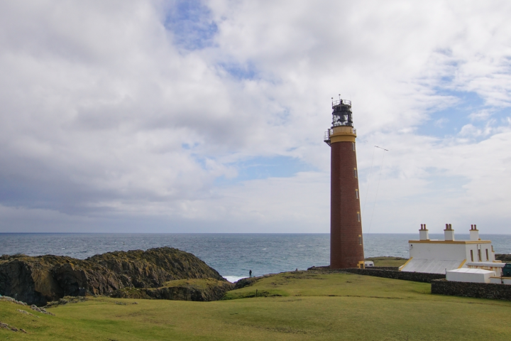 Tall brick lighthouse on grassy coast with cloudy sky and ocean.