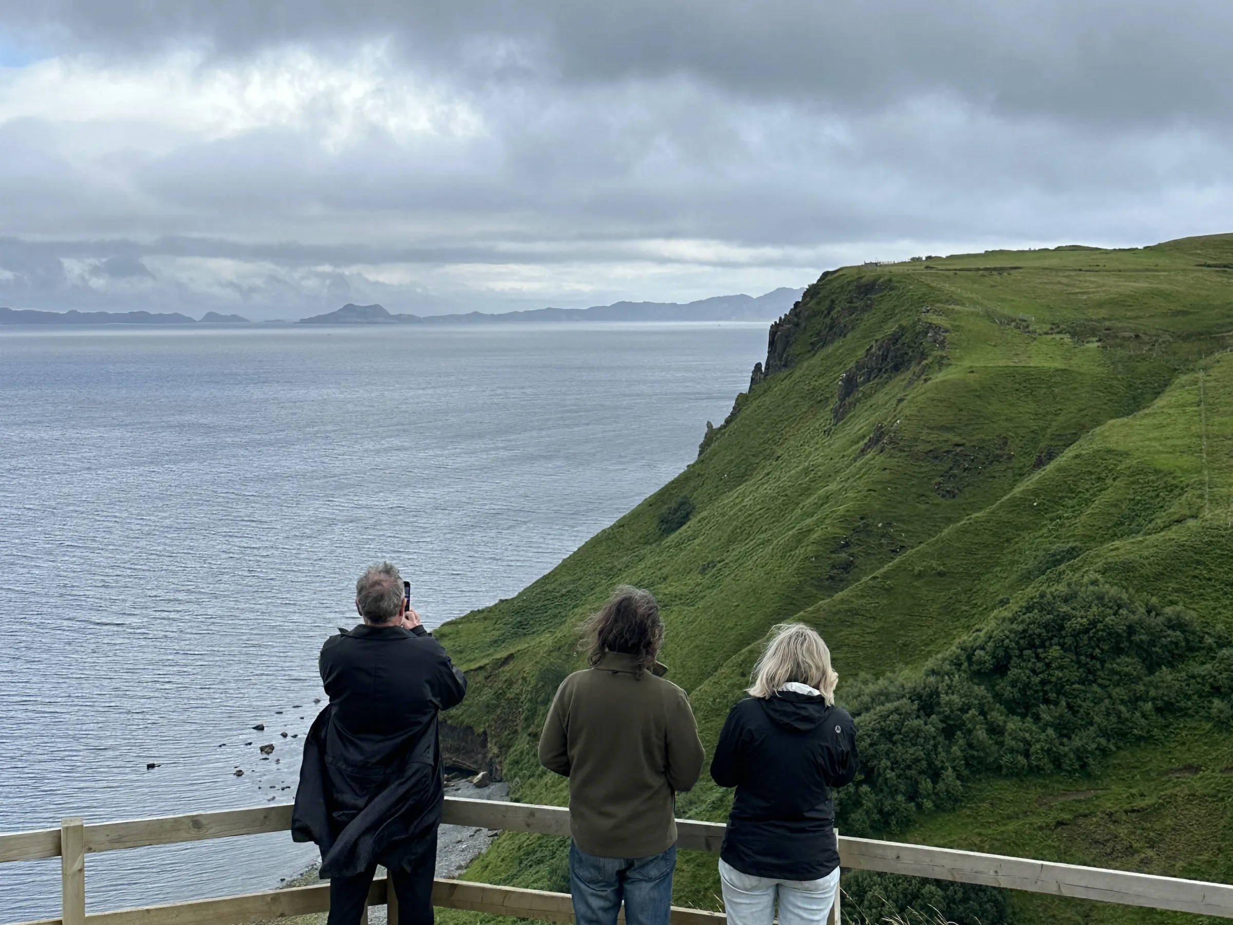 Three people at a cliff edge with a view of the sea and distant hills.