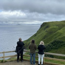 Three people at a cliff edge with a view of the sea and distant hills.