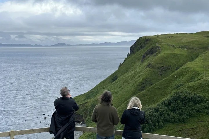 Three people at a cliff edge with a view of the sea and distant hills.