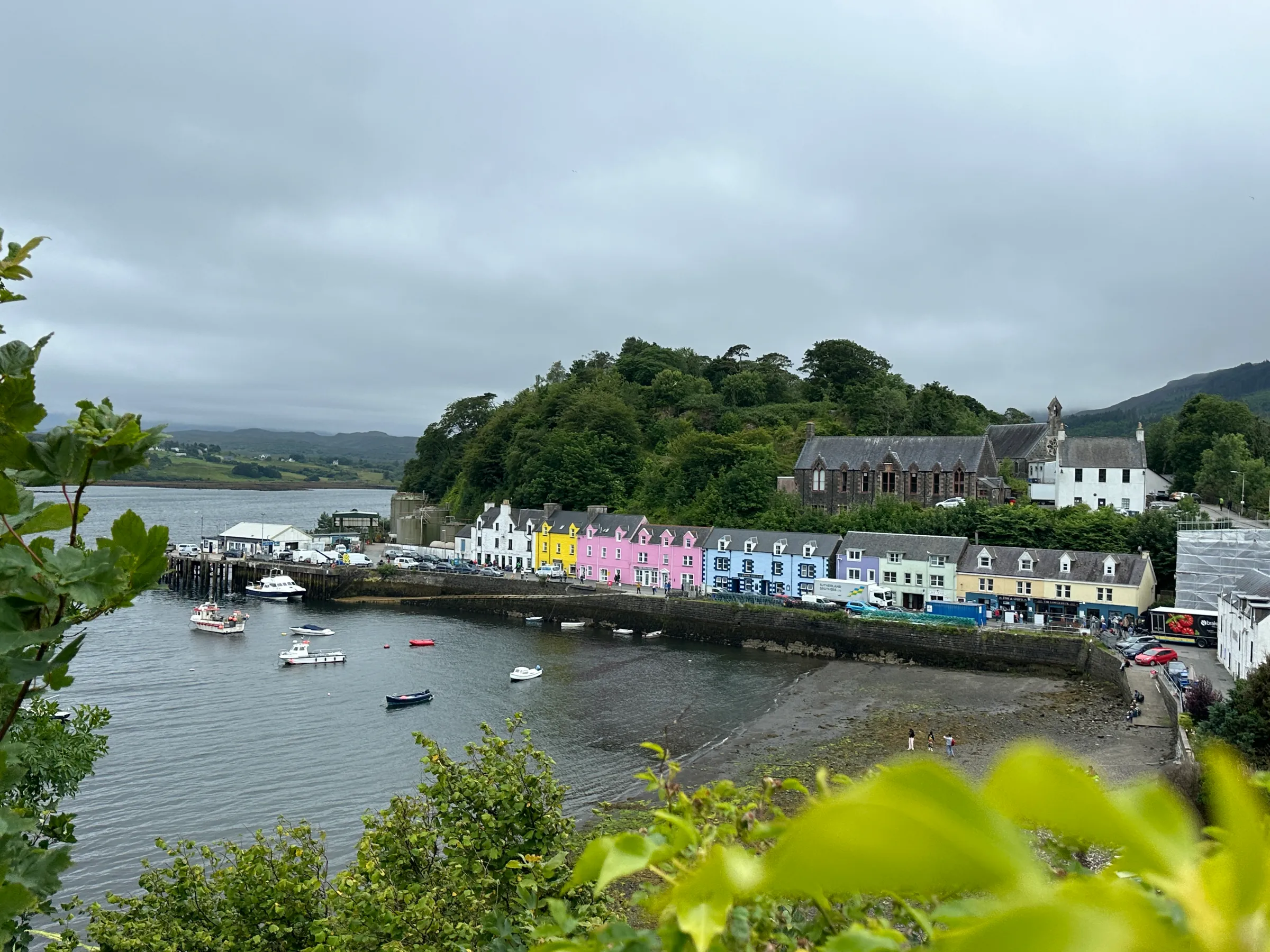 Colorful harborfront buildings with boats on a cloudy day, surrounded by hills and greenery.