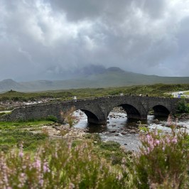 Stone bridge over a river in a green landscape with cloudy sky in the background.