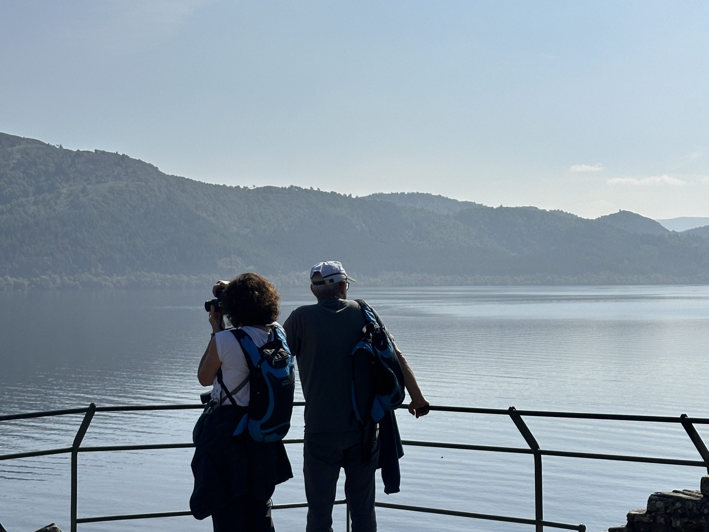 Two people with backpacks overlooking a lake from a railing on a clear day.