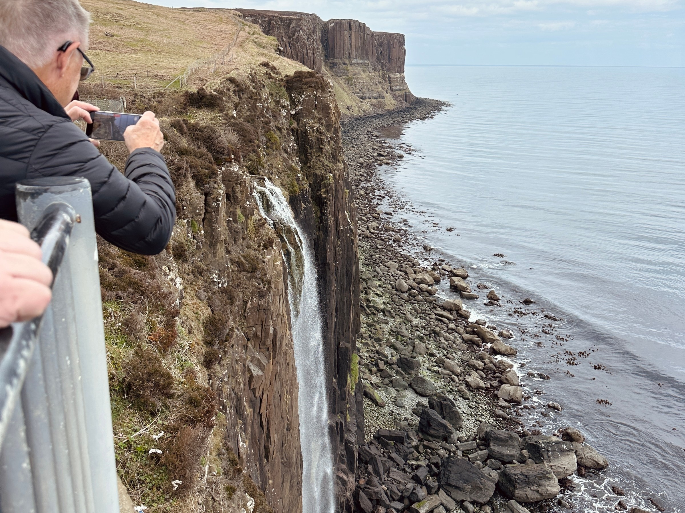 Person photographing a cliffside waterfall overlooking a rocky coastline.