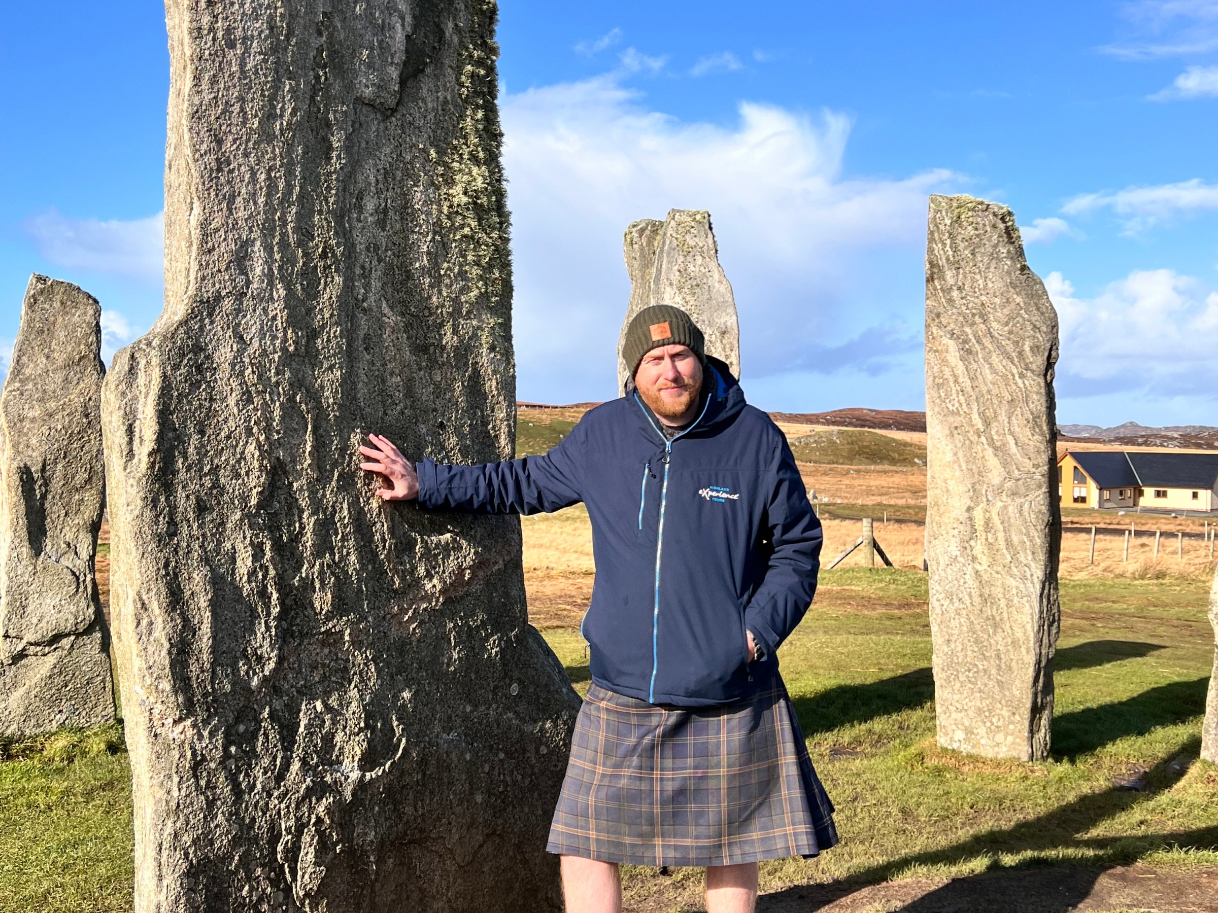 Man in a kilt stands by tall stone pillars on a sunny day.