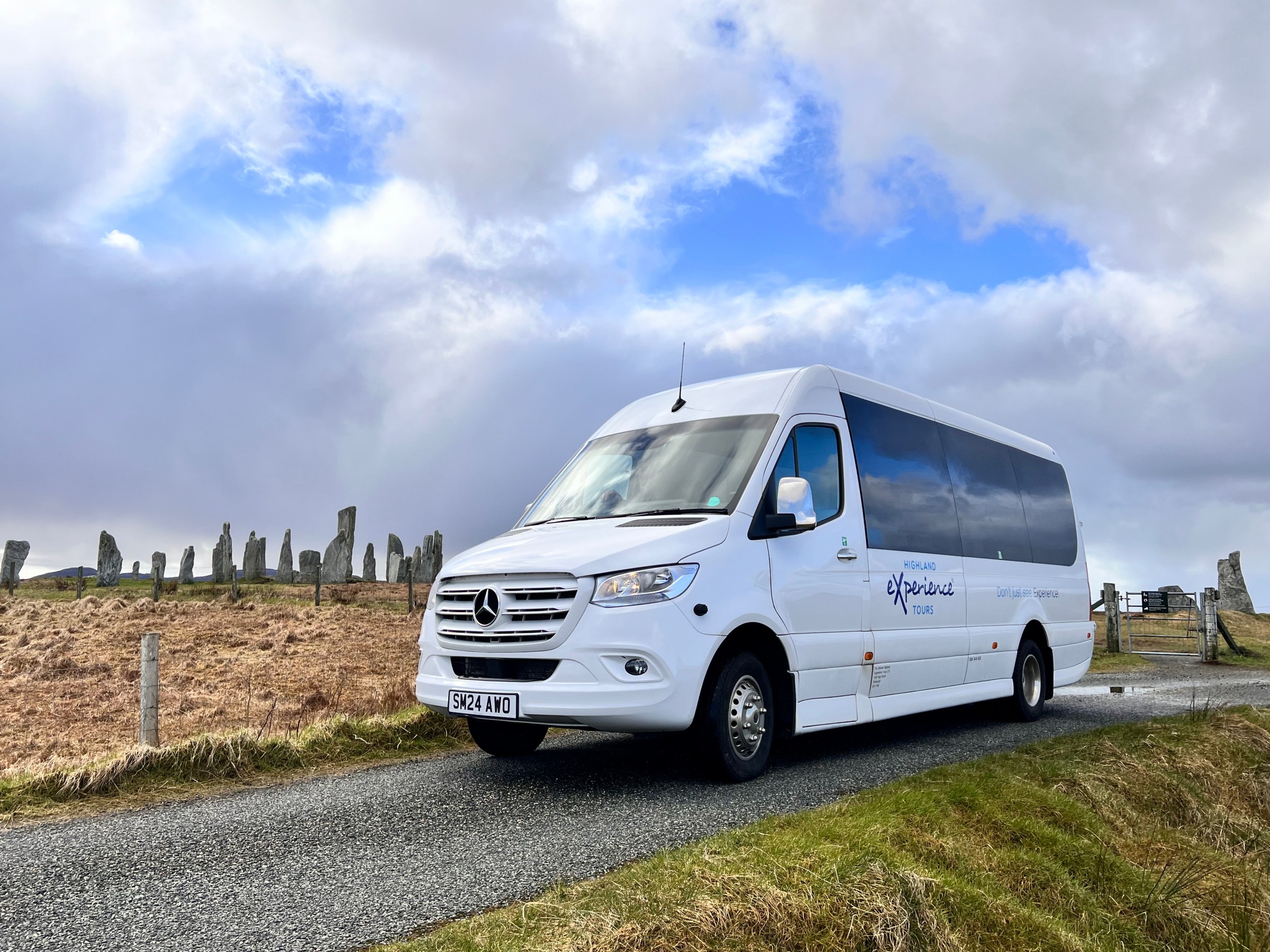 White van on rural road with stone structures in the background under cloudy sky.