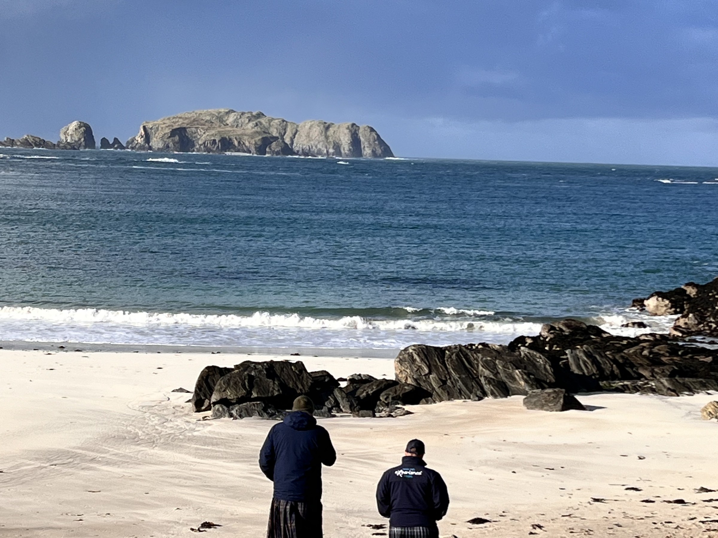 Two people in kilts walking towards a rocky beach under a cloudy sky.