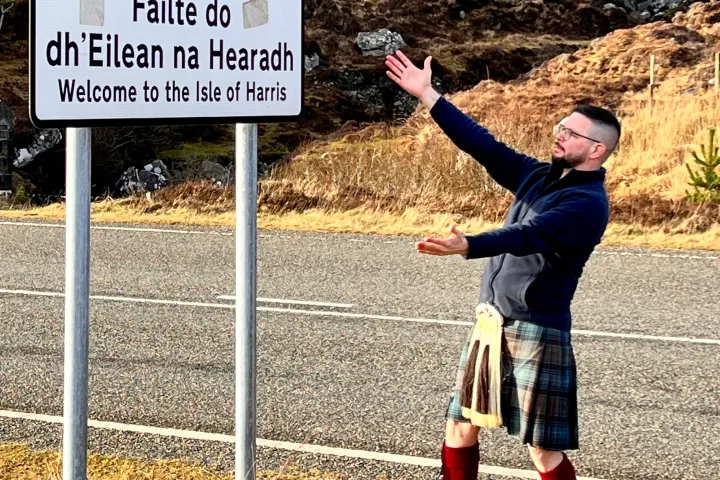Person in kilt gesturing towards Isle of Harris welcome sign.