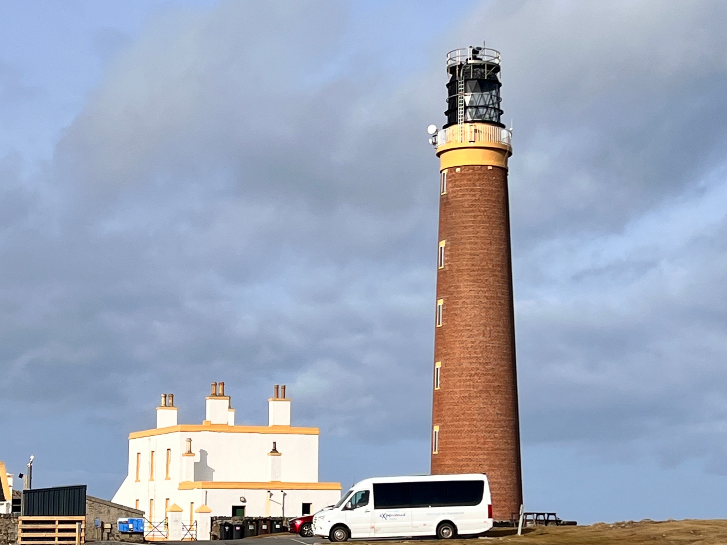 Tall brick lighthouse with white building and van on a cloudy day.