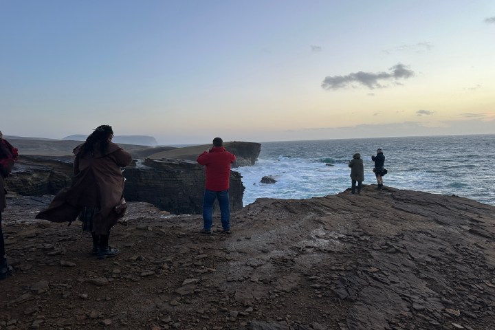 People on a rocky cliff overlooking the ocean at sunset.
