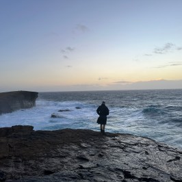 Person standing on rocky shore with ocean waves at sunset.