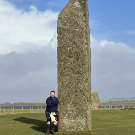 Person standing beside a large, upright stone monolith on a grassy field.