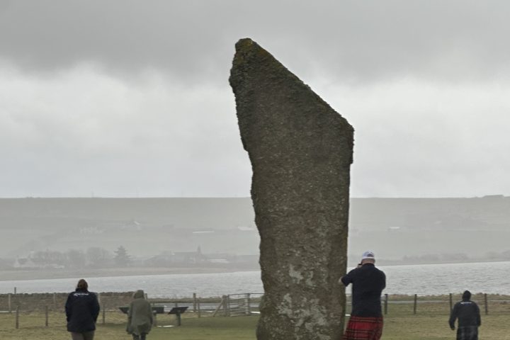 People in kilts near a standing stone on a grassy field by the sea.