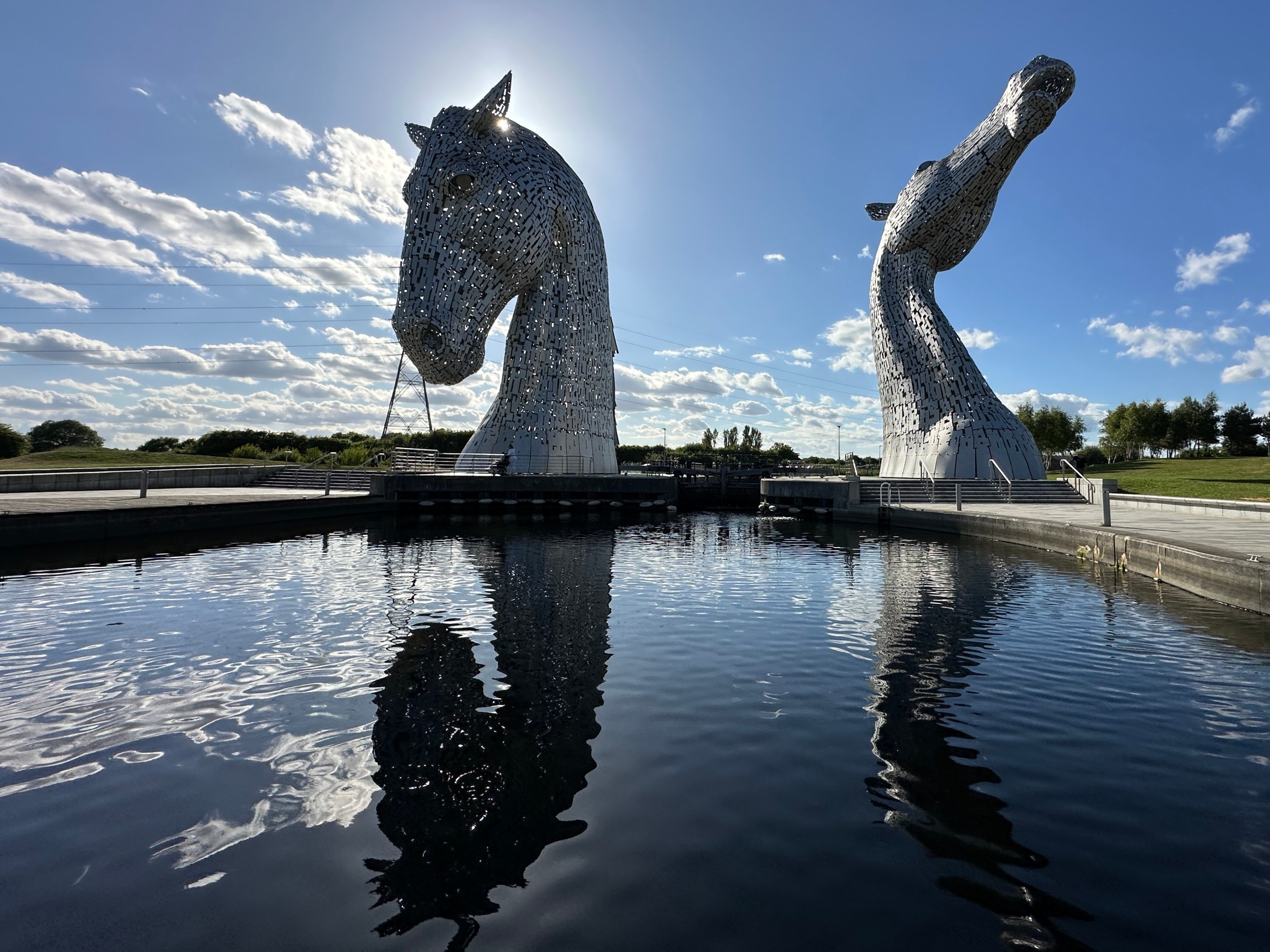 Two large horse head sculptures reflected in a water channel under a blue sky.