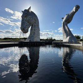 Two large horse head sculptures reflected in a water channel under a blue sky.