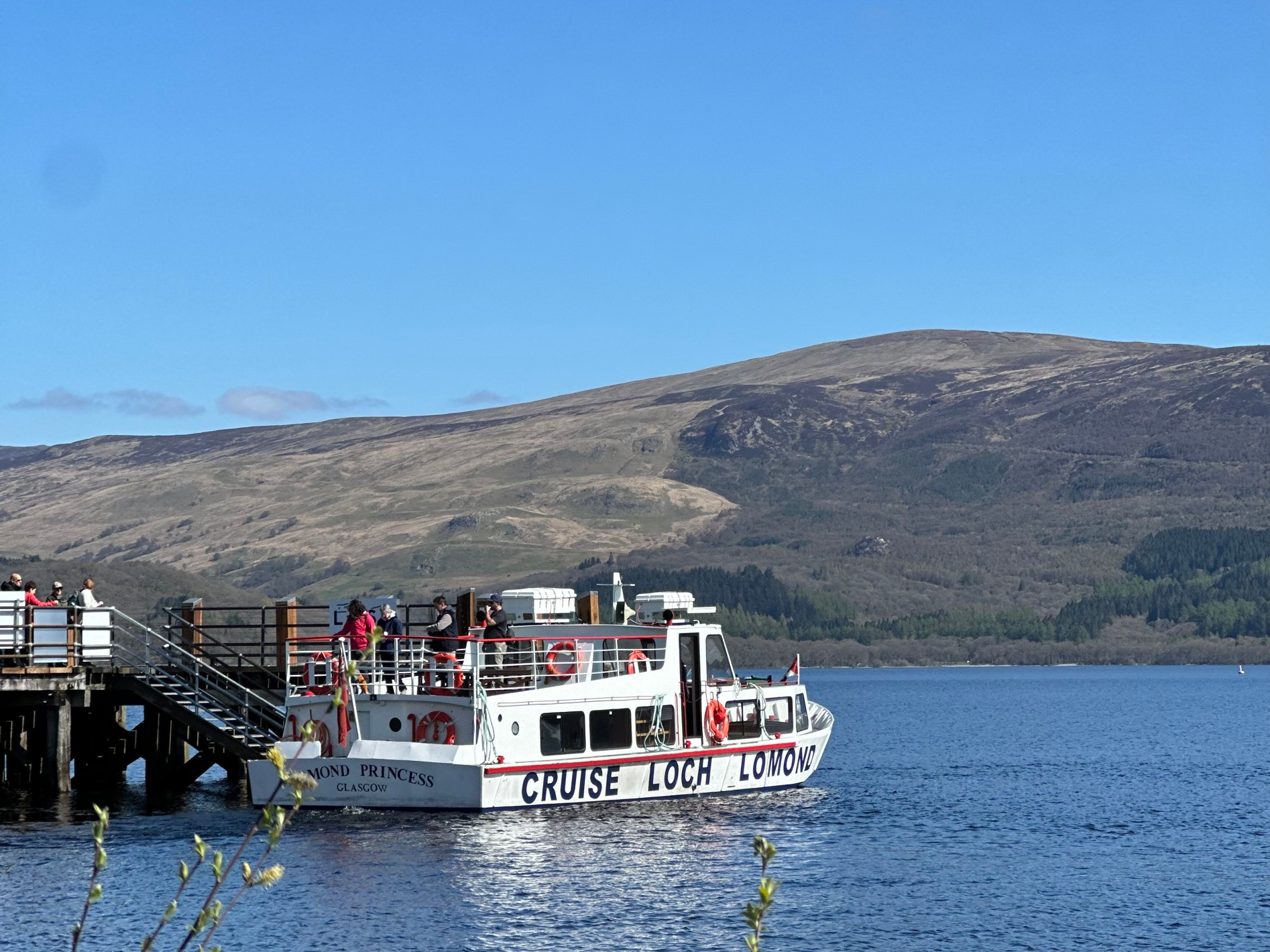 Tour boat docked at Loch Lomond with hills in the background under a clear blue sky.