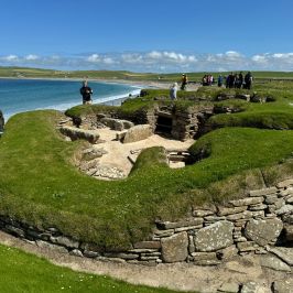 People exploring ancient ruins on a grassy coastal site under a clear blue sky.