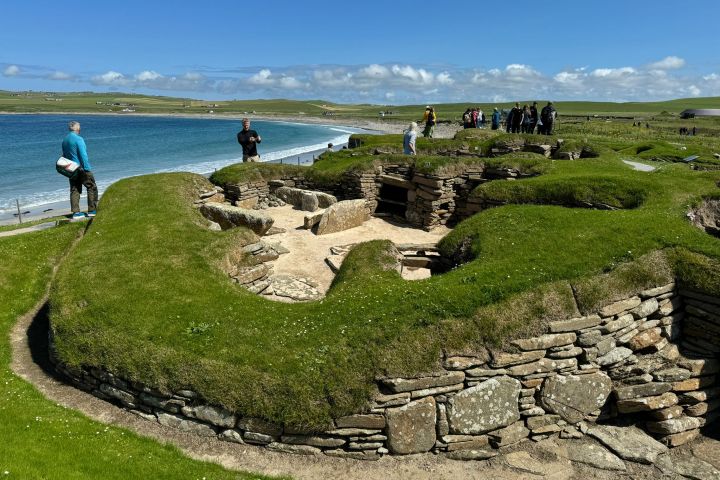 People exploring ancient ruins on a grassy coastal site under a clear blue sky.