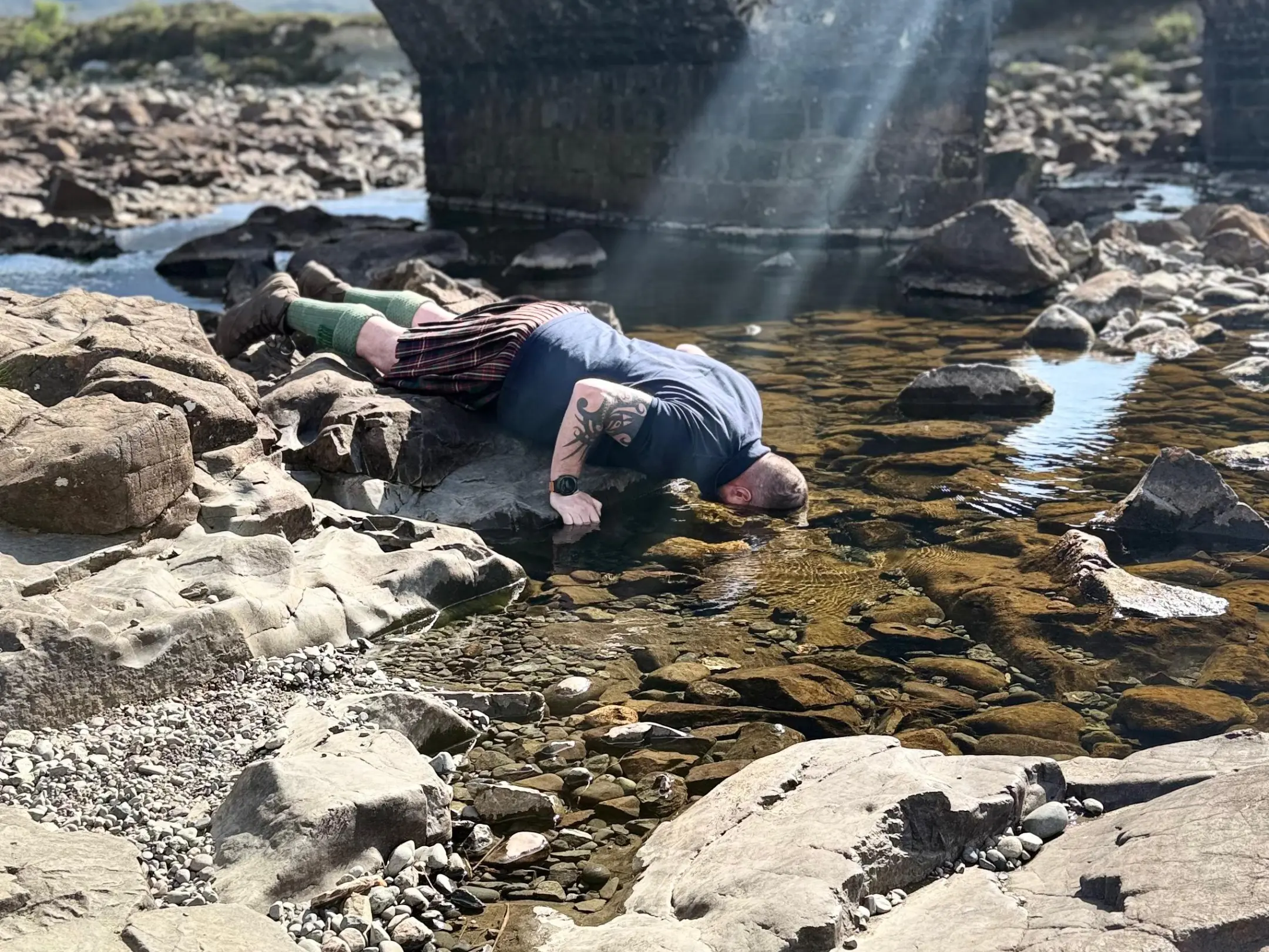 Person drinking from a stream under a stone bridge with sunlight streaming down.