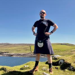 Man in kilt standing on grassy hill by a lake on a sunny day.