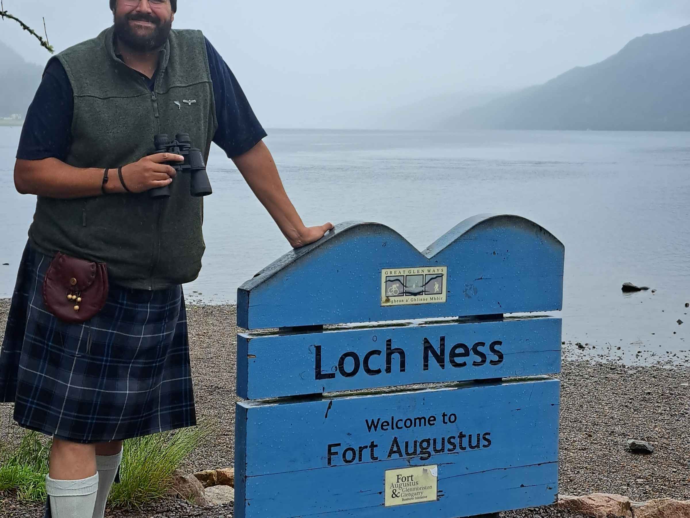 Man in kilt stands by Loch Ness sign with binoculars.