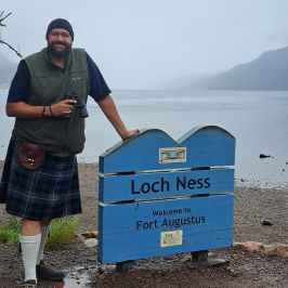 Man in kilt with binoculars stands by Loch Ness sign near water.