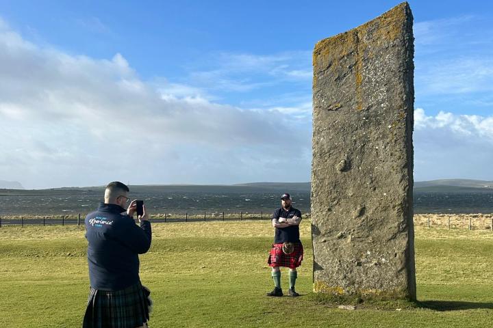 Two men in kilts by a large standing stone in a grassy field.