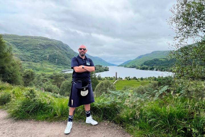 Man in kilt stands on hillside, overlooking a lake and mountains.