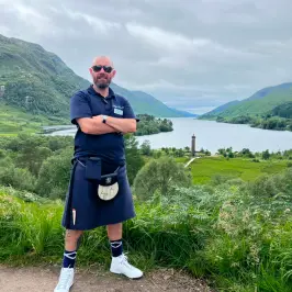 Man in kilt stands with arms crossed, mountains and lake in background.