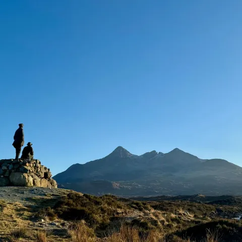 Two people on a rock pile in front of mountain landscape under clear blue sky.