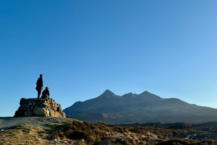 Two people on a rock pile in front of mountain landscape under clear blue sky.
