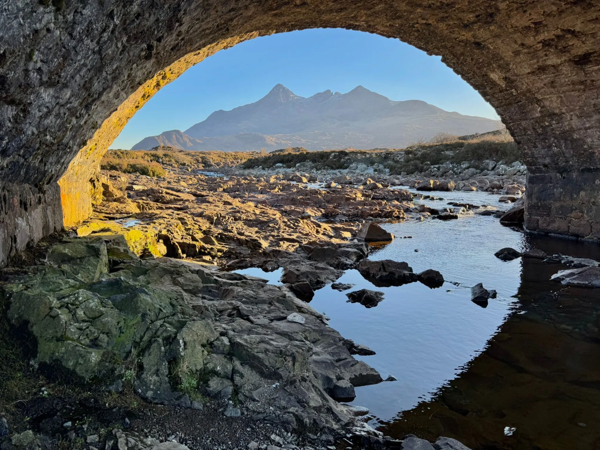 Rocky stream view through stone arch with distant mountain landscape.