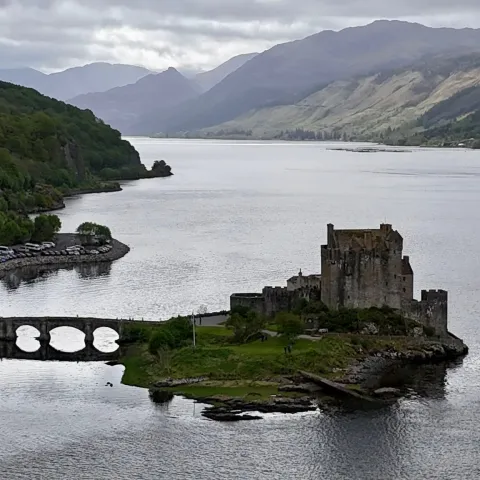 Stone castle on a lake with surrounding hills under a cloudy sky.