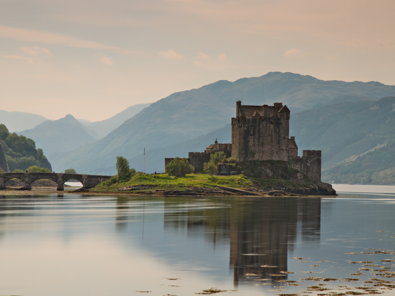 A castle on an island with a bridge in the foreground and mountains in the background.
