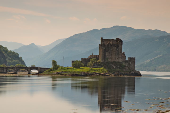A castle on an island with a bridge in the foreground and mountains in the background.