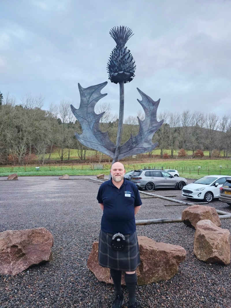 Man in kilt stands before large thistle sculpture in a parking lot with cars and trees in the background.