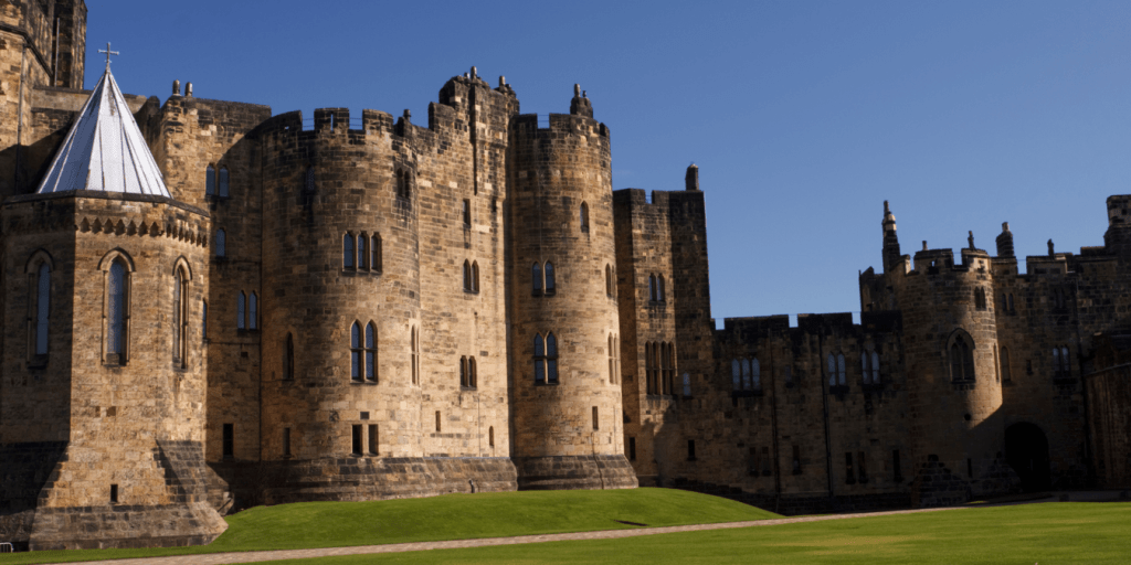 a stone castle with a clock tower in front of a brick building with Alnwick Castle in the background