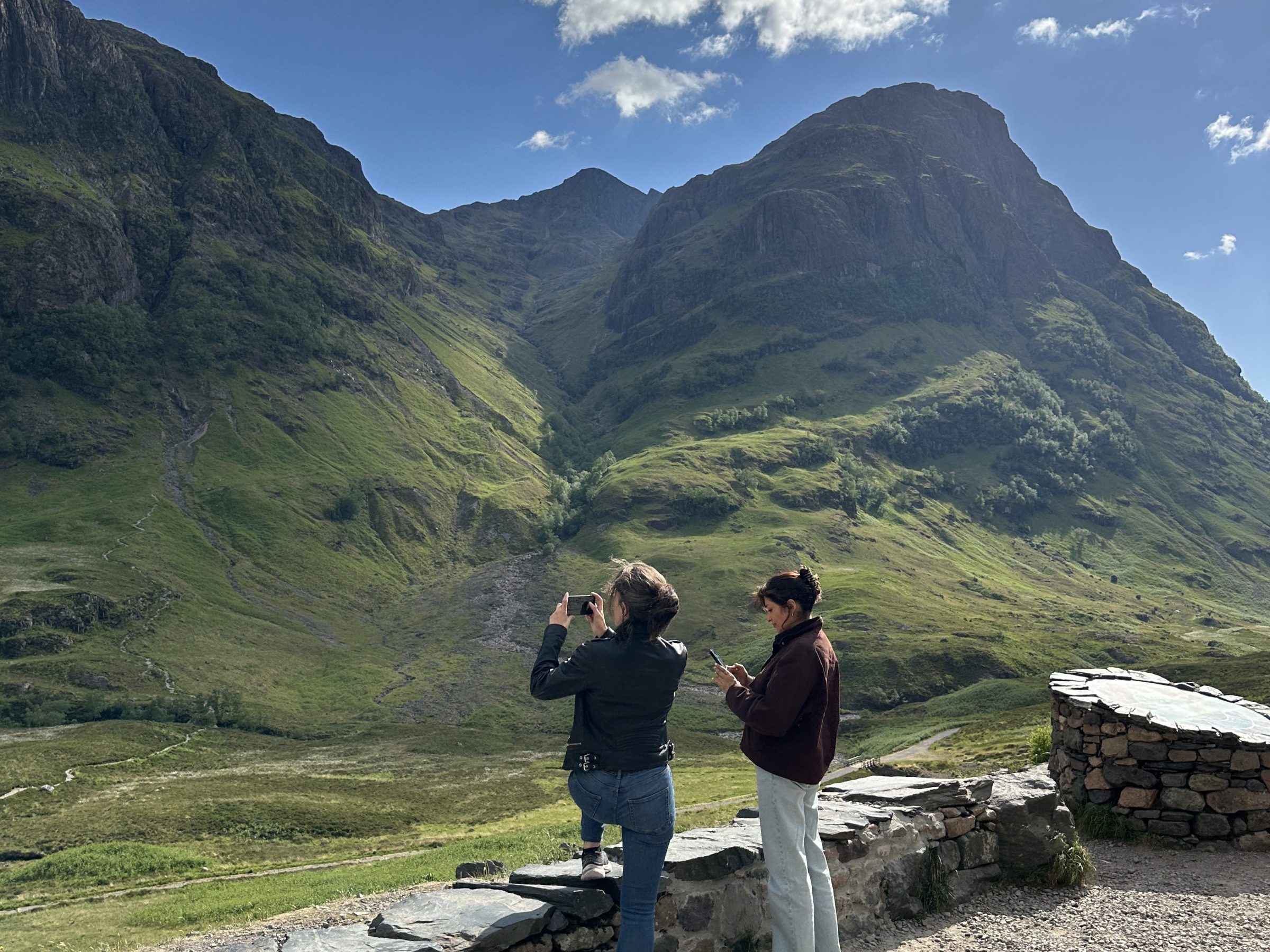 Two people taking photos of a mountainous landscape under a cloudy blue sky.