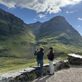 Two people taking photos of a mountainous landscape under a cloudy blue sky.
