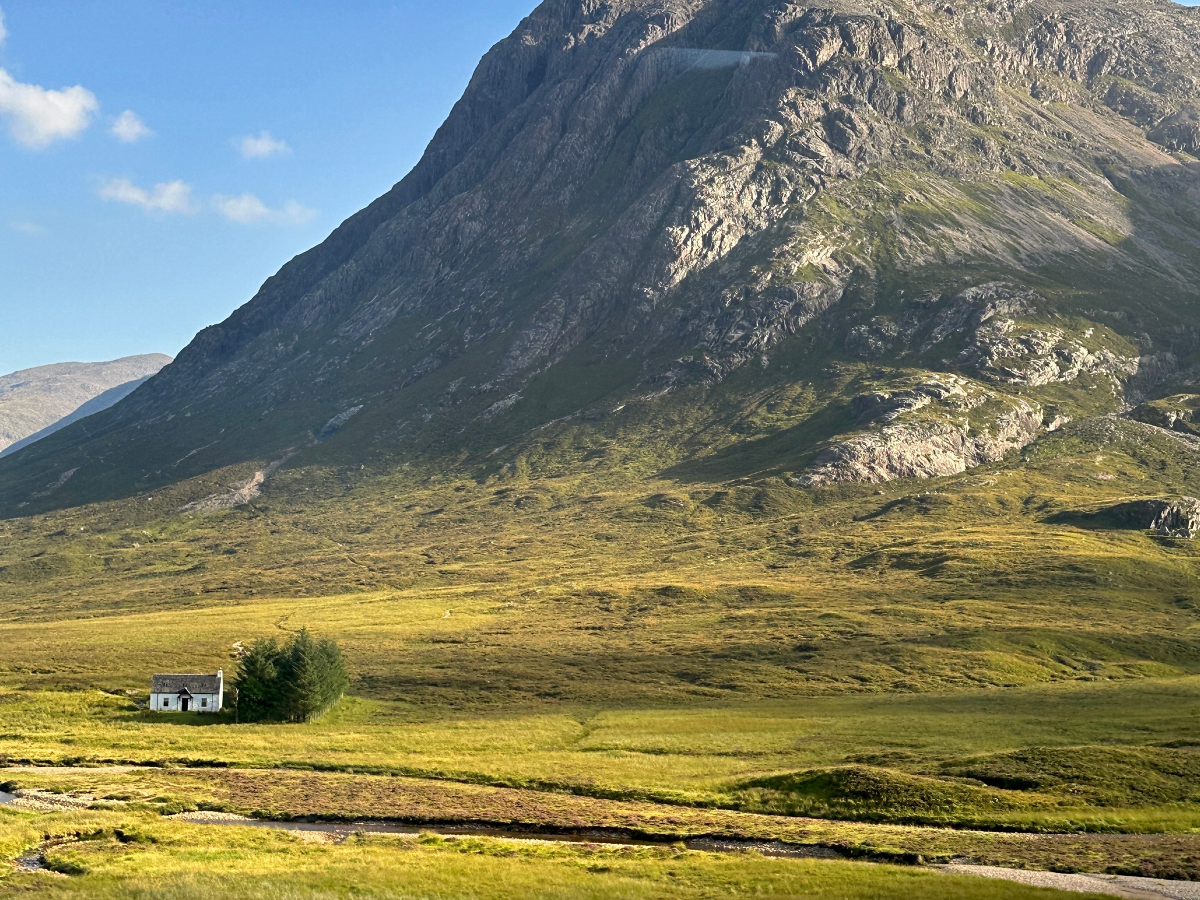 Mountain landscape with a small white house and grassy field under a blue sky.