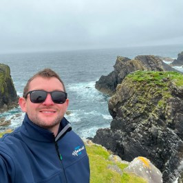 Person in sunglasses and jacket smiles near rocky coastline and cloudy sky.