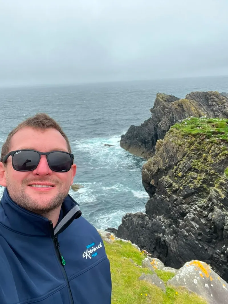 Person in sunglasses and jacket smiles near rocky coastline and cloudy sky.