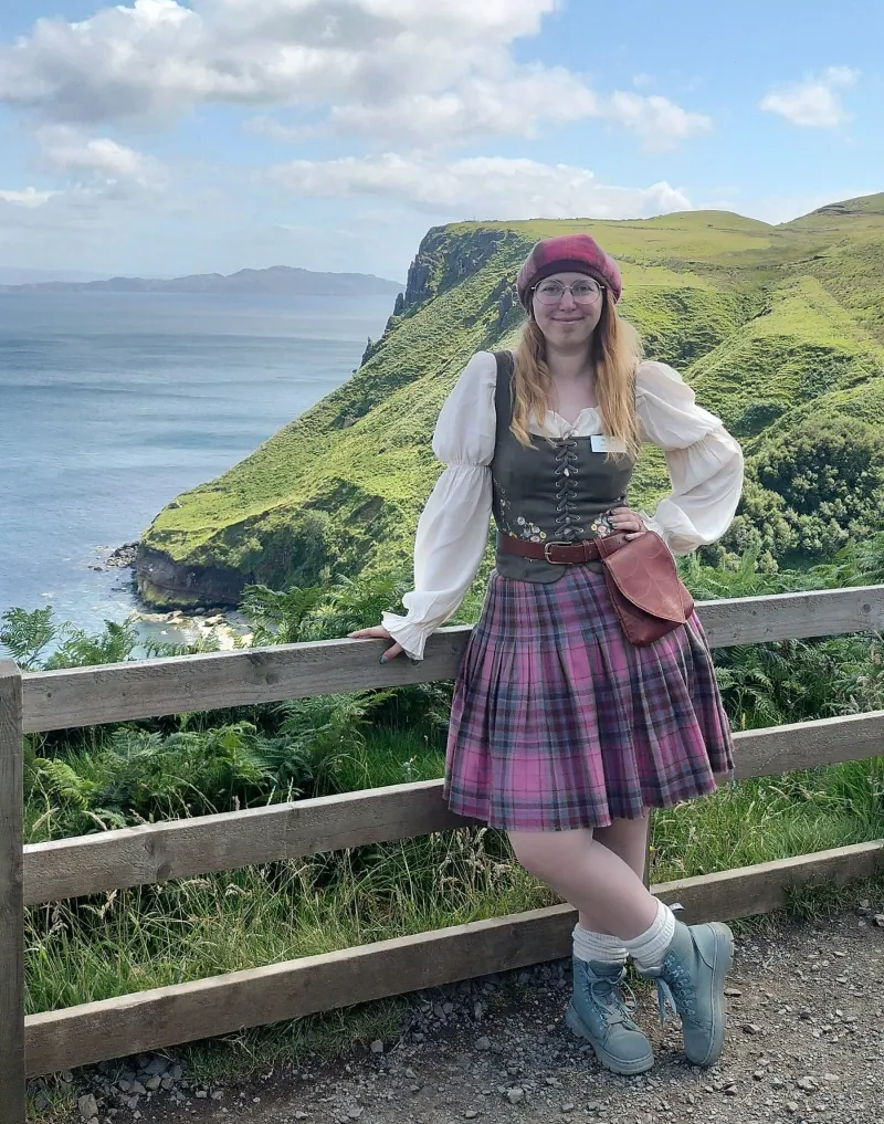 Person in traditional attire standing by fence with hillside and sea in background.